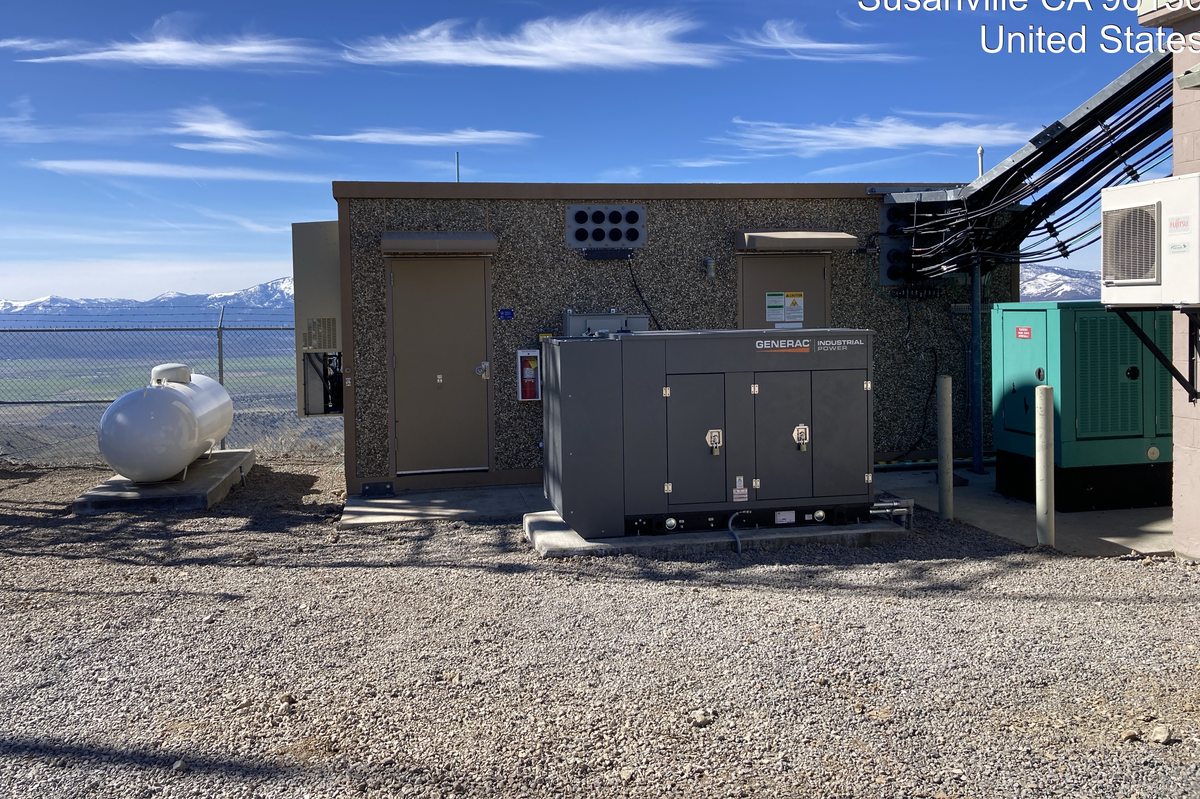 Generator and equipment shelter at a Susanville mountain site with snow-capped peaks behind